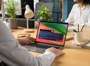 A person seated at a desk in an office setting is using a laptop displaying a colorful interface with a vibrant gradient background. The screen shows the Ava platform.