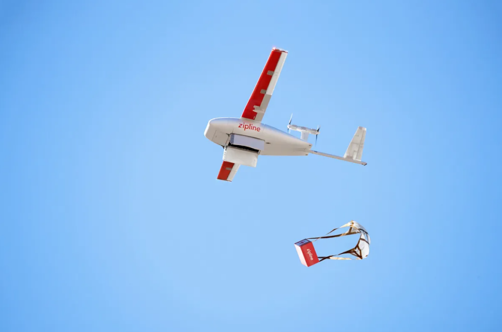 A Zipline drone flying in the sky, releasing a package of medicine mid-air for delivery.