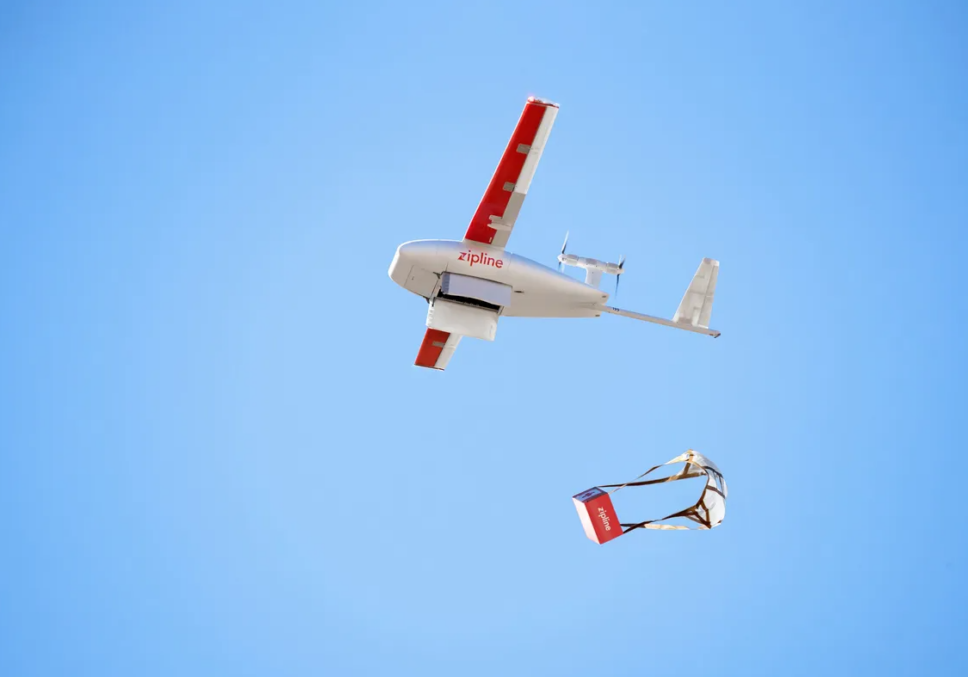 A Zipline drone flying in the sky, releasing a package of medicine mid-air for delivery.